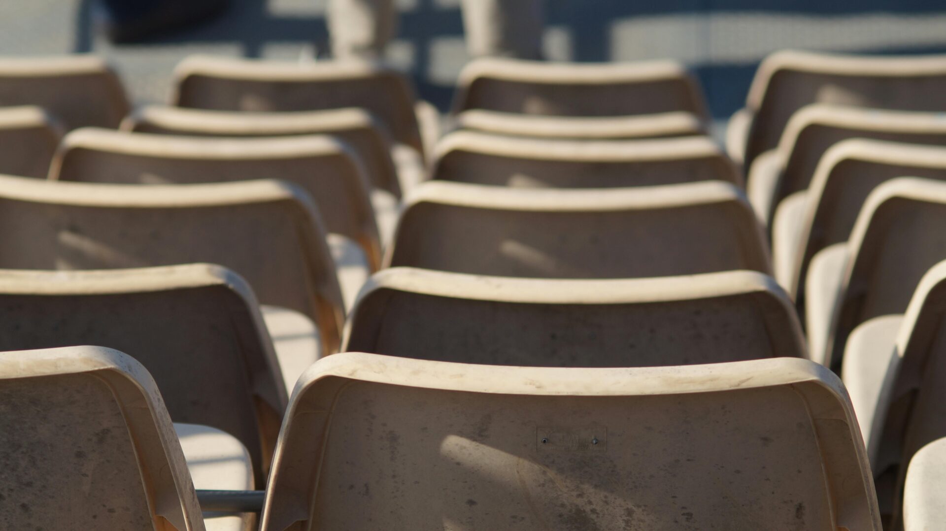 Empty rows of plastic backrest chairs in an outdoor stadium under sunlight.